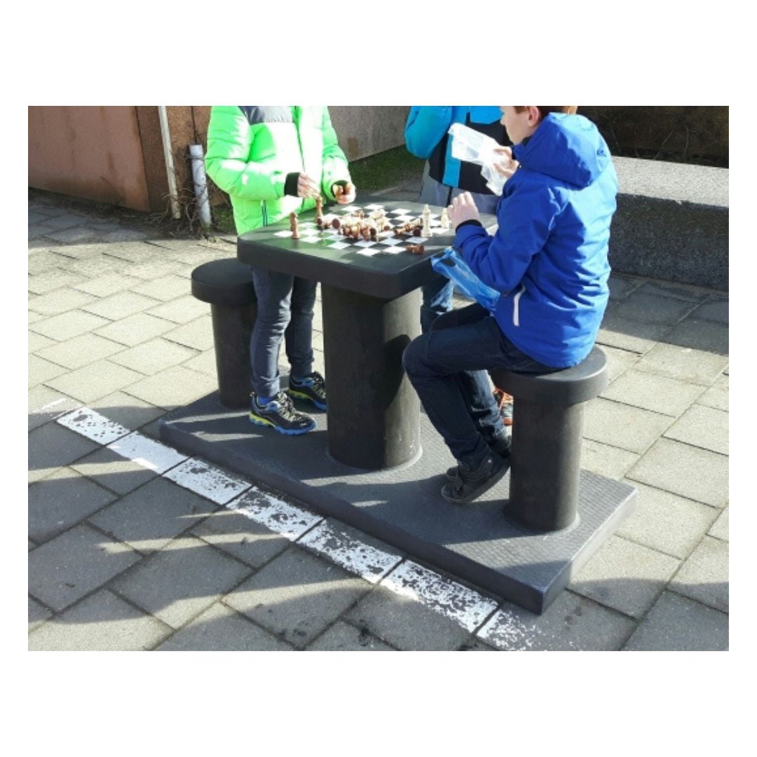 to børn sidder ved et skakbord i beton og spiller skak i en indkørsel med en tilskuer i blå jakke