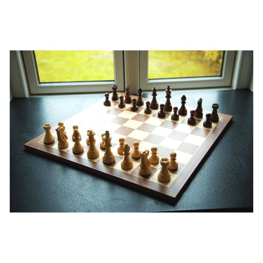 Skakspil i træ stående på en mørk flade i en vindues karm ud til en have, hvor lyset står ind og lader skakspillet kaste skygger af sig